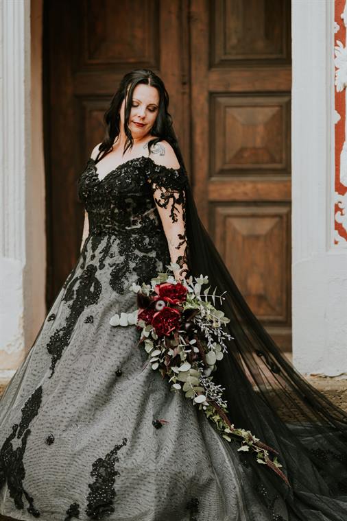 Braut im schwarzen Kleid mit dunkelrotem Strauß vor einer Holztür